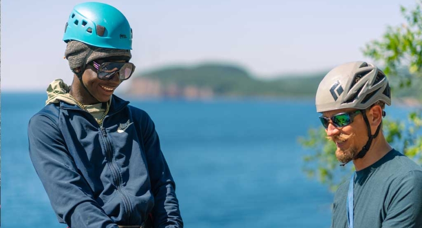 Two people wearing helmets and sunglasses smile. They appear to be at a high elevation. There is water below them.